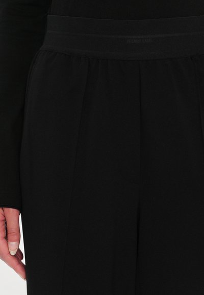 Pantaloni neri con una texture liscia e una vita ampia. Presentano delle pieghe sulla parte anteriore e un logo discreto all'altezza della vita.