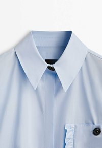 Chemise bleu clair avec un col pointu, une poche avant ornée d'un détail à volants et un bouton décoratif noir sur la poche. Texture lisse.