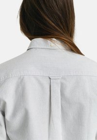 Donna con capelli castani che indossa una camicia bianca con sottili righe verticali nere, vista di schiena con focus sul colletto e sulla parte superiore della schiena.