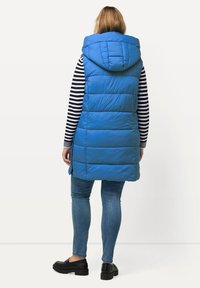 Blå puffväst med huva, med horisontell sömnad, bärs över en svart och vit randig långärmad skjorta, ihop med jeans och svarta loafers.
