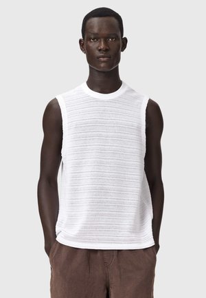 Homme debout, regardant vers l'avant, portant un haut tricoté blanc sans manches et un pantalon marron, les mains dans les poches, sur un fond gris.