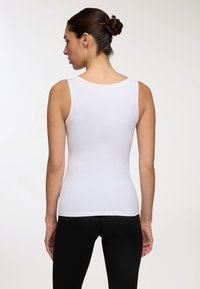 Donna con capelli scuri raccolti a chignon, vestita con una maglietta bianca aderente senza maniche e pantaloni neri, vista di spalle su uno sfondo semplice.