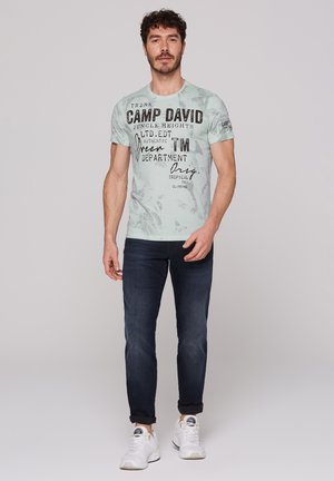 Light mint green short-sleeve t-shirt with black printed text and abstract patterns, paired with dark blue jeans and white sneakers.