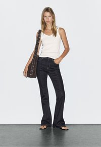 Hellbeige Tanktop, dunkle lässige Jeans mit taillierter Passform und braune Schultertasche mit Nieten, getragen mit schwarzen Flip-Flops.