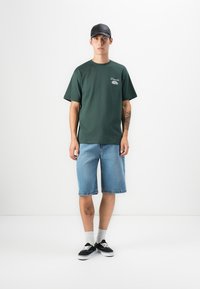 Green short-sleeve t-shirt with white logo, light blue denim shorts, patterned socks, and black sneakers with white soles. Black cap worn.