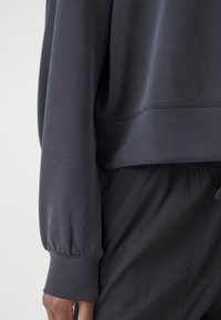 Sötét szürke, hosszú ujjú pulóver, begyűrt mandzsettákkal és kerek szegéllyel, sötét, texturált nadrággal párosítva. Puha, sima anyag.