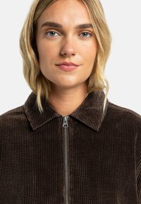 Brown corduroy jacket with a collared neckline and zipper closure. Features vertical ribbing texture and a fitted design.