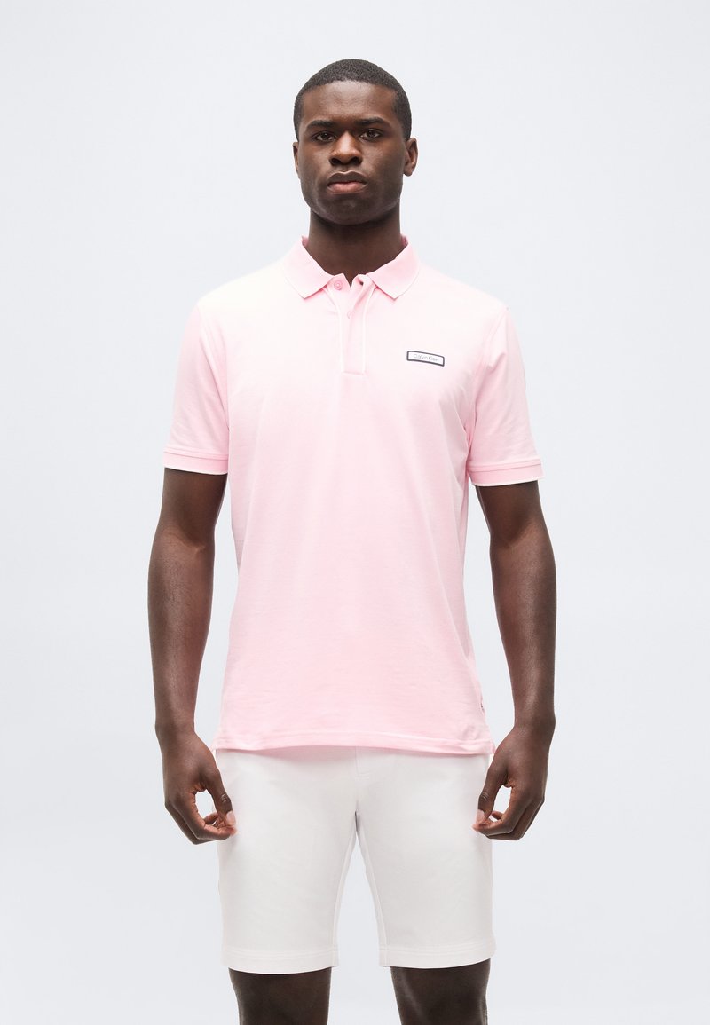 Homme portant un polo rose clair et un short blanc, debout devant un fond blanc uni, face à l'avant.