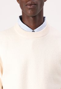 Pull en maille couleur crème avec un col rond côtelé, superposé à une chemise à col rayée bleu clair et blanche. Texture douce.