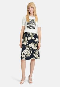 Witte T-shirt met korte mouwen en grafische tekst, gecombineerd met een marineblauwe midi-rok met bloemen, voorzien van crème en groene bladeren, afgemaakt met witte blokhakken.