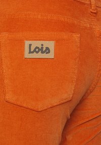 Oranžová manšestrová tkanina s čtvercovou zadní kapsou, na které je tančí kožená nášivka s vyraženým názvem značky "Lois". Viditelná texturovaná plocha a švy.