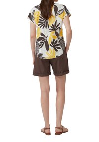 Chemise à manches courtes avec une base blanche et des motifs de feuilles jaune vif et noir, associée à un short marron, retroussé à l'ourlet, et des sandales marron.