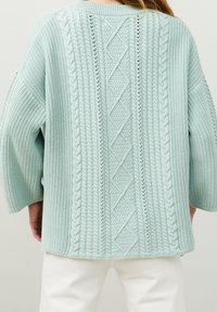 Femme portant un pull en maille texturée bleu clair avec des motifs torsadés et en diamant, associé à un pantalon blanc, vue de dos.