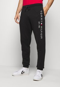 Mann trägt schwarze Tommy Hilfiger Sweatpants mit Kordelzug in der Taille und weiße Sneakers, steht vor einem schlichten hellen Hintergrund.