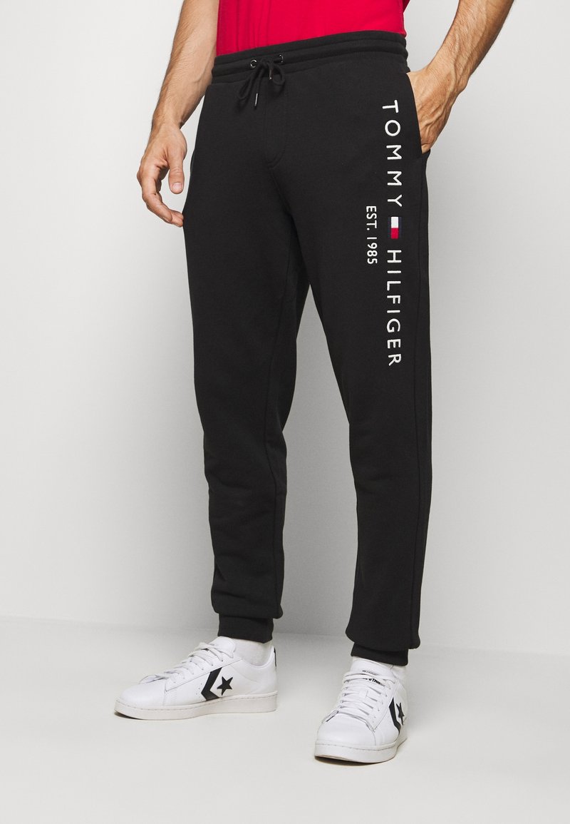 Mann trägt schwarze Tommy Hilfiger Sweatpants mit Kordelzug in der Taille und weiße Sneakers, steht vor einem schlichten hellen Hintergrund.