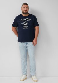 Granatowy bawełniany T-shirt z białym nadrukiem graficznym i tekstem. W połączeniu z jasnoniebieskimi dżinsami i białymi trampkami. Prosty, casualowy fason.