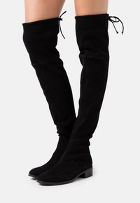 Bottes en suède noires au-dessus du genou avec un bout arrondi, un petit talon carré et des liens réglables en haut pour un maintien sécurisé.