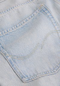 Το ανοιχτό μπλε τζιν ύφασμα διαθέτει πίσω τσέπη με καμπυλωτή ραφή και κεντήματα σε κίτρινο χρώμα που ξεχωρίζουν, παρουσιάζοντας μια διακριτική υφή και σχεδιασμό.