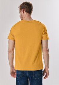 Žluté tričko s krátkým rukávem a kulatým výstřihem; hladký bavlněný materiál; viditelné švy na ramenech a rovný lem; džíny.