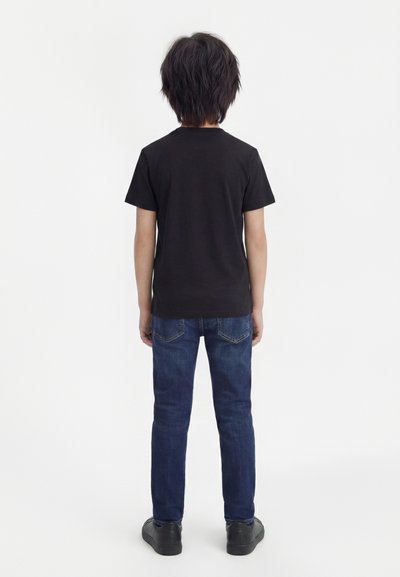 Παιδί που στέκεται με την πλάτη γυρισμένη, φοράει ένα απλό μαύρο κοντομάνικο πουκάμισο, μπλε τζιν και μαύρα παπούτσια σε λευκό φόντο.