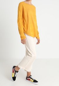 Persona che indossa un maglione lungo a maniche lunghe di un giallo senape con texture, pantaloni a vita alta con orlo sfrangiato di colore crema e scarpe da ginnastica nere con design di fiamme rosse e gialle.