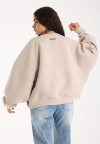 Kobieta z długimi ciemnymi włosami, ubrana w oversize'owy beżowy sweter i jasnoniebieskie dżinsy, pokazana z tyłu na tle gładkiej białej ściany.