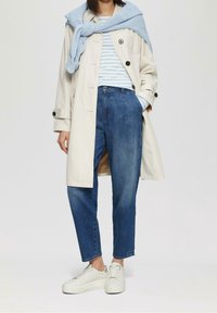 Osoba v béžovém trenčkotě, modrém proužkovaném tričku, modrých džínách, bílých teniskách a světlomodrém svetru přehozeném přes ramena.