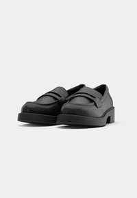 Chaussures en cuir noir avec un bout arrondi, une semelle plate et un détail de sangle sur le cou-de-pied. Texture lisse avec des accents de couture minimaux.