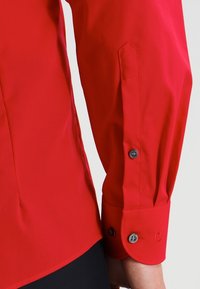 Rotes Langarmhemd mit glatter Textur, Manschetten mit dunklen Knöpfen und taillierter Passform. Vorgeformter Ärmel mit leicht zulaufender Form.