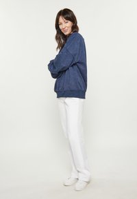 Tengerészkék pulóver, laza illeszkedéssel és bordázott mandzsettákkal, fehér egyenes szárú nadrággal és fehér sportcipővel párosítva.