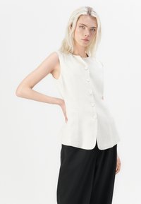 Cream sleeveless blouse with a high round neckline, featuring five front buttons, a fitted bodice, and side pockets. Paired with black pants.