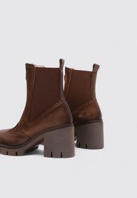 Botines marrones hasta el tobillo con una combinación de materiales lisos y texturizados. Tacones gruesos en bloque y angulados con paneles laterales elásticos para facilitar su uso.