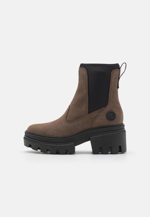 Bottine en cuir marron avec panneaux latéraux extensibles noirs, semelle crantée épaisse, talon bloc et languette à l’arrière.