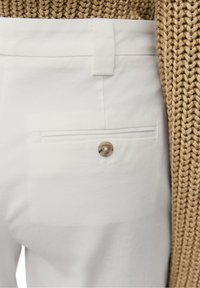 Pantalon blanc avec une poche arrière unique dotée d'un bouton rond de couleur claire. Le tissu semble lisse avec une coupe structurée.
