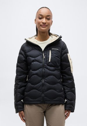 Žena oblečená v čiernej prešívanej bunde s béžovými detailmi, predným zipsom a vreckami na zips, stojaca na jednoliatom bielom pozadí.