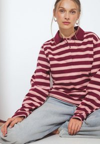 Giovane donna seduta, che indossa una polo a maniche lunghe a righe bordeaux e rosa e jeans azzurri, con orecchini a cerchio e capelli raccolti.