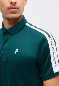 Gros plan d'un homme portant un polo vert foncé avec des rayures blanches et noires sur l'épaule et le texte "Peak Performance" sur la manche.