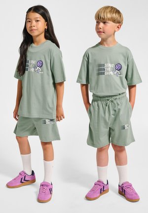 Lichtgroene katoenen T-shirts met een 'Serve the Game'-grafiek, gecombineerd met bijpassende shorts. Schoeisel is een paar roze sneakers met witte sokken.