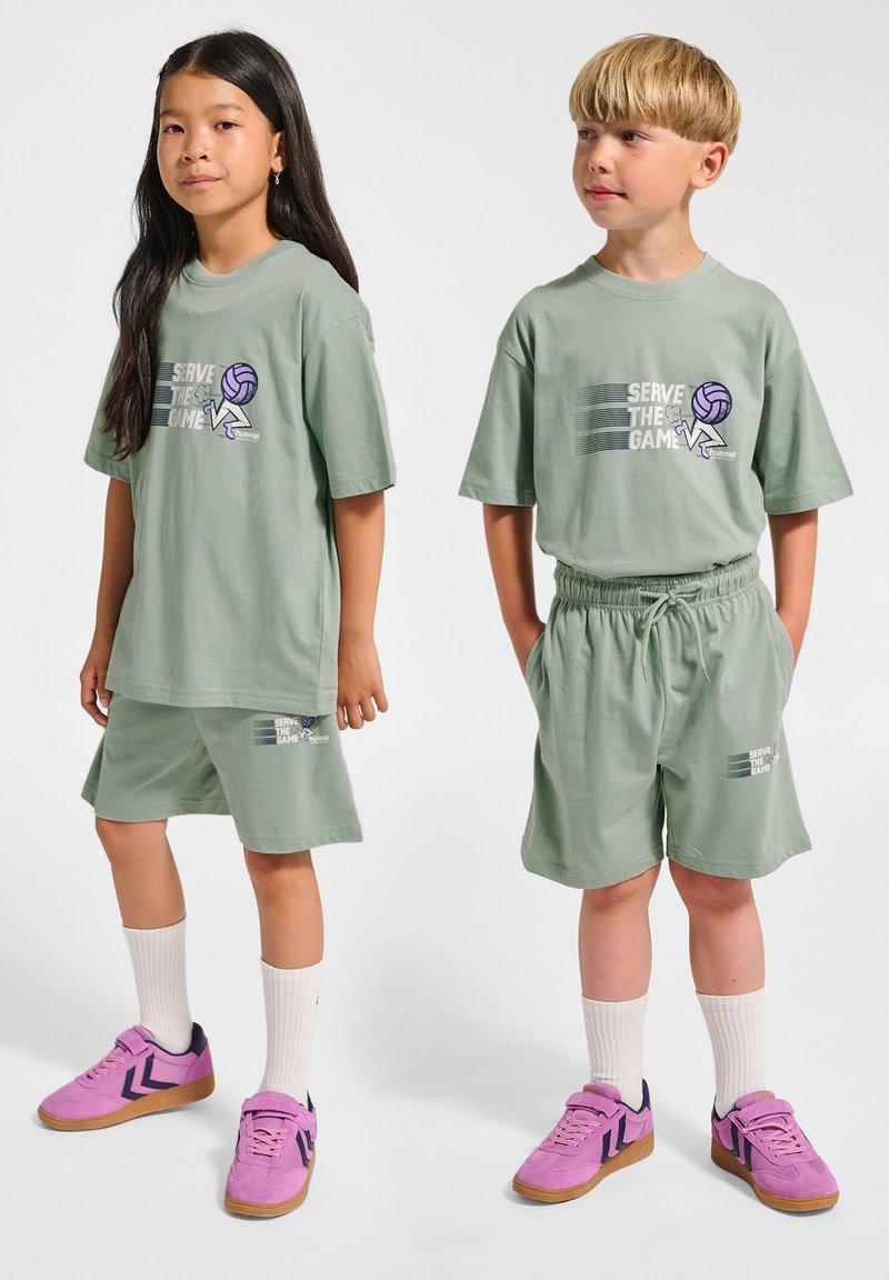 Ljusgröna bomulls-T-shirts med grafik som säger 'Serve the Game', matchade med byxor i samma färg. Skorna är rosa sneakers med vita strumpor.