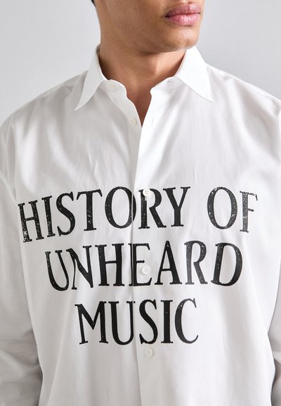 Vit skjorta med knappar och svart tryckt text "SÄLLSYNT MUSIKS HISTORIA". Slät tyg med klassisk krage och normal passform.