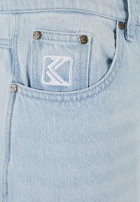 Un bolsillo de jeans de mezclilla azul claro con un logotipo geométrico bordado en blanco y cuatro remaches metálicos con detalles del logotipo a juego.