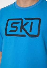 Sininen puuvillapaita, jossa on iso musta "SKI" -logo edessä. Pyöreä kaula-aukko ja lyhyt hiha, tasainen ja miellyttävä pinta.