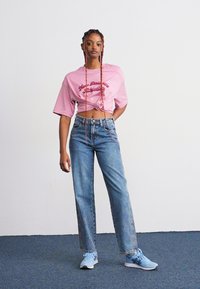 Jeune femme avec de longs cheveux tressés se tient debout, portant un t-shirt graphique rose court, un jean bleu taille haute et des baskets bleu clair sur un tapis bleu.