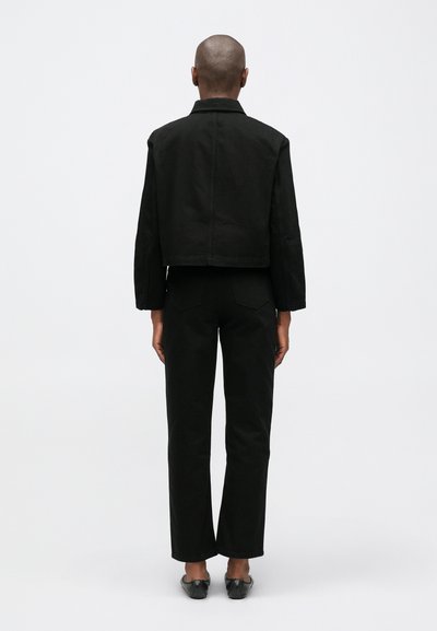 Lühemate juustega inimene kannab musta lühikest jakki, musti sirgeid pükse ja musti libisevaid jalatseid, seistes seljaga tavalise tausta vastu.