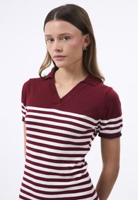 Polo a maniche corte a righe rosse e bianche con colletto a V e design aderente. I dettagli includono rifiniture blu sui maniche.