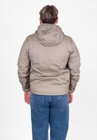 Giacca imbottita marrone chiaro con cappuccio, caratterizzata da una texture liscia e lucida e polsini elastici. Indossata sopra jeans di denim blu.