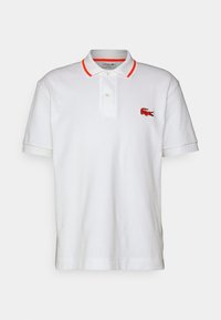 Polo shirt blanc en coton avec un col classique, deux boutons et des accents orange sur le col, présentant un logo de crocodile brodé en rouge.