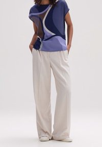 Person klädd i en kortärmad topp med abstrakt mönster i marinblått, lavendel och brunt, tillsammans med vida beige byxor och vita skor, med händerna i fickorna.