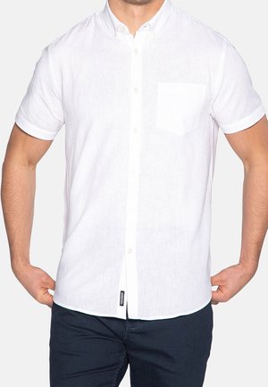 Homme portant une chemise blanche à manches courtes avec une poche poitrine et un pantalon sombre, les mains reposant près des hanches.