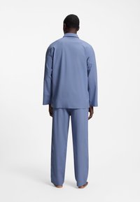 Άνδρας που στέκεται ξυπόλητος με την πλάτη προς το φακό, φορώντας χαλαρό ανοιχτό μπλε μακρυμάνικο πάνω και παντελόνι στον ίδιο τόνο, σε λευκό φόντο.